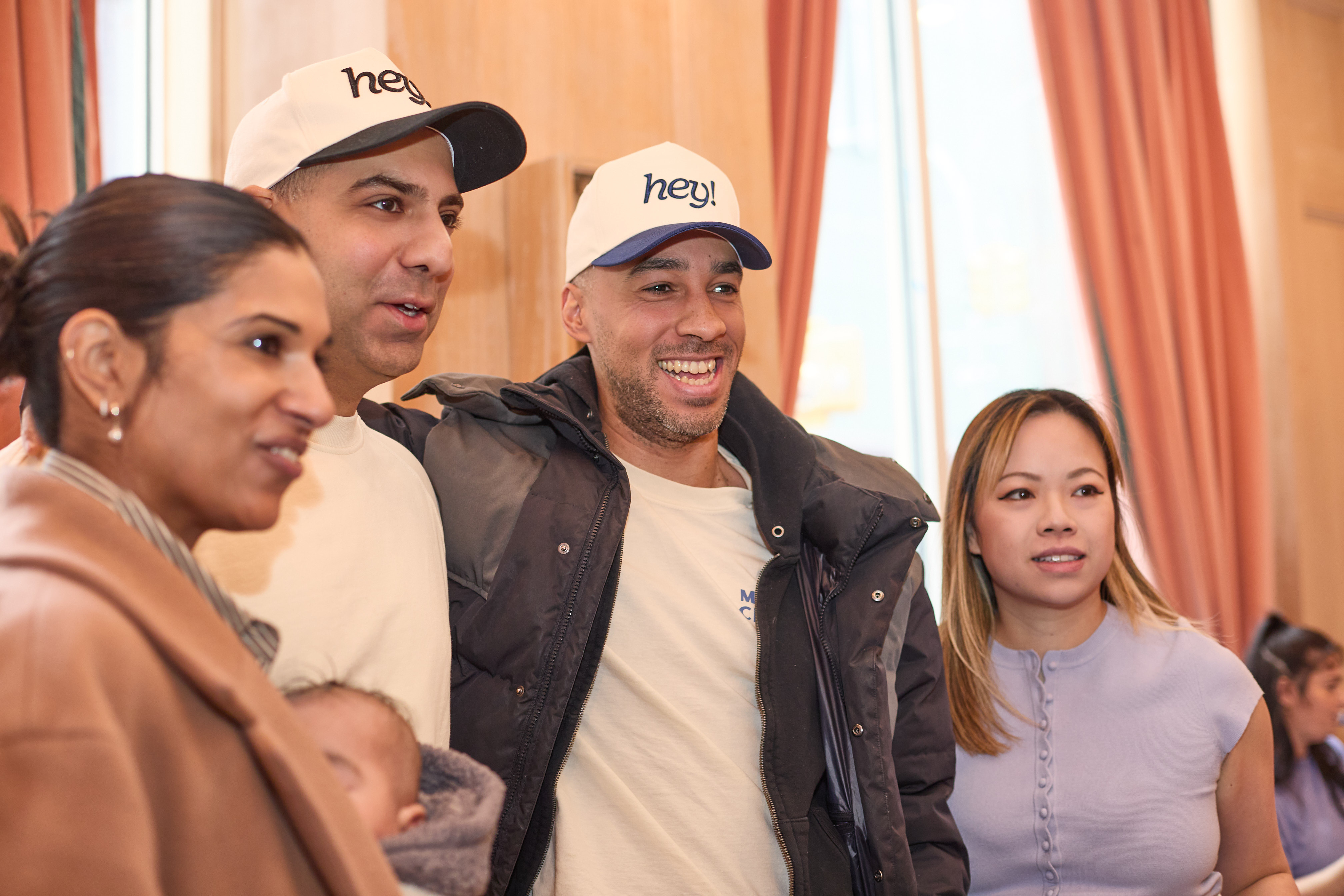 Two men wearing 'hey!' hats sitting next to two women in a casual setting.