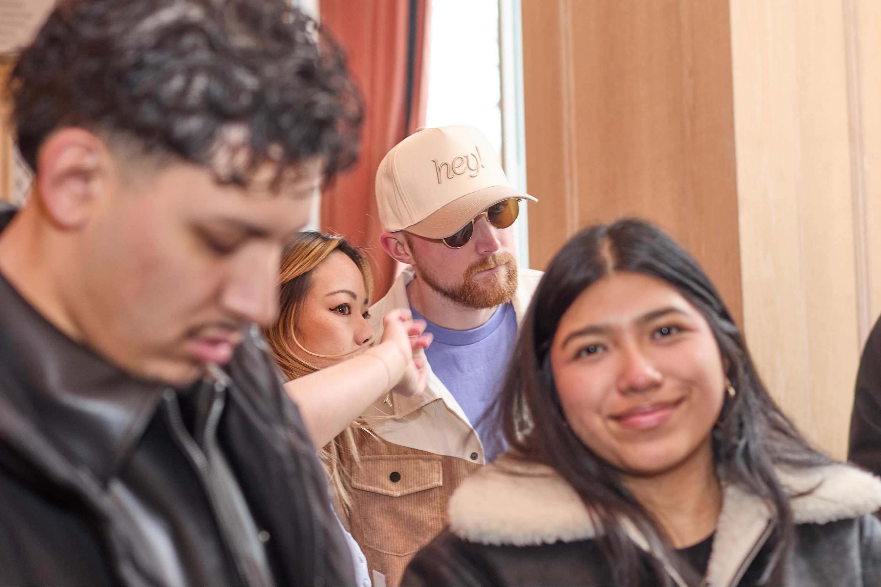 Group of people standing indoors with a wooden door and framed picture in the background. A guy wearing a beige hey hat in the middle.