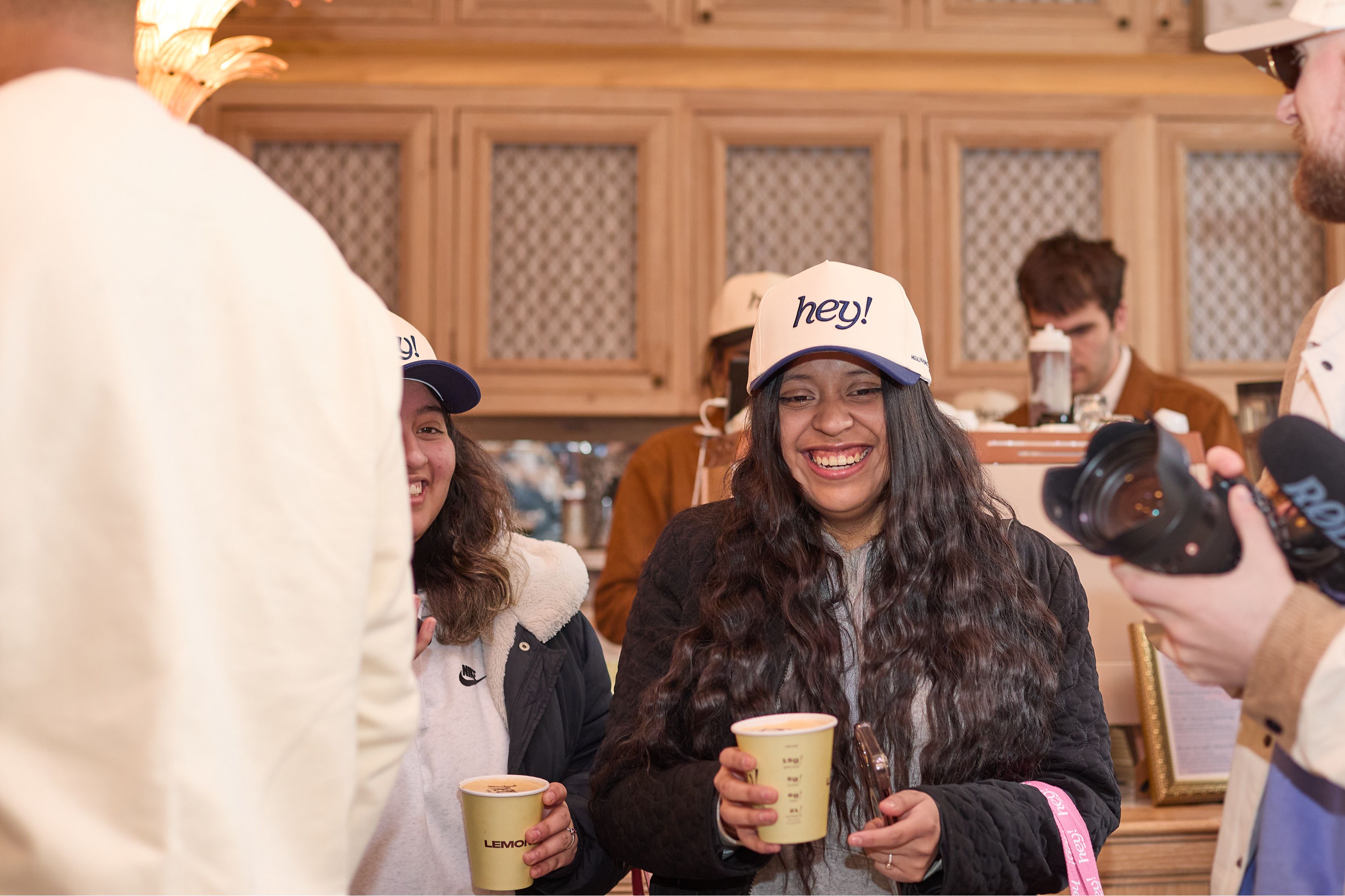 Smiling woman wearing a “hey!” baseball cap holds a yellow drink cup while chatting in a cafe; a photographer with a camera stands nearby and others in matching hats are in the background.