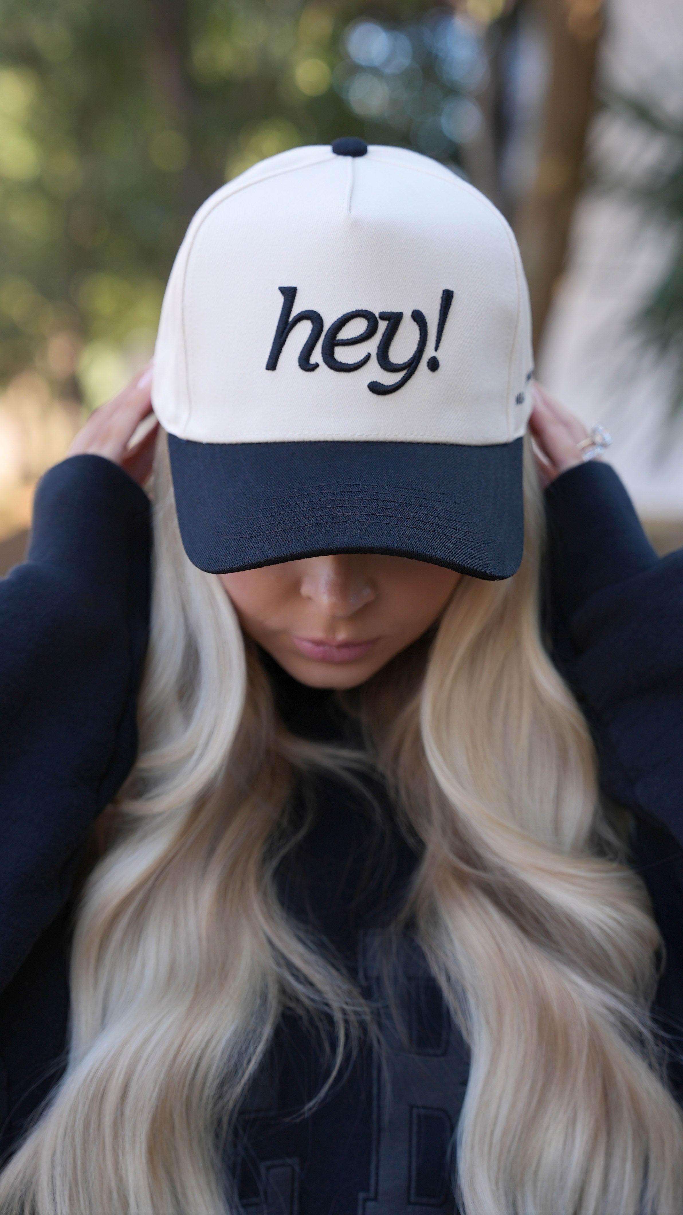 Kay Dudley wearing a cream “hey!” embroidered hat outdoors, smiling with her head tilted slightly to the side.