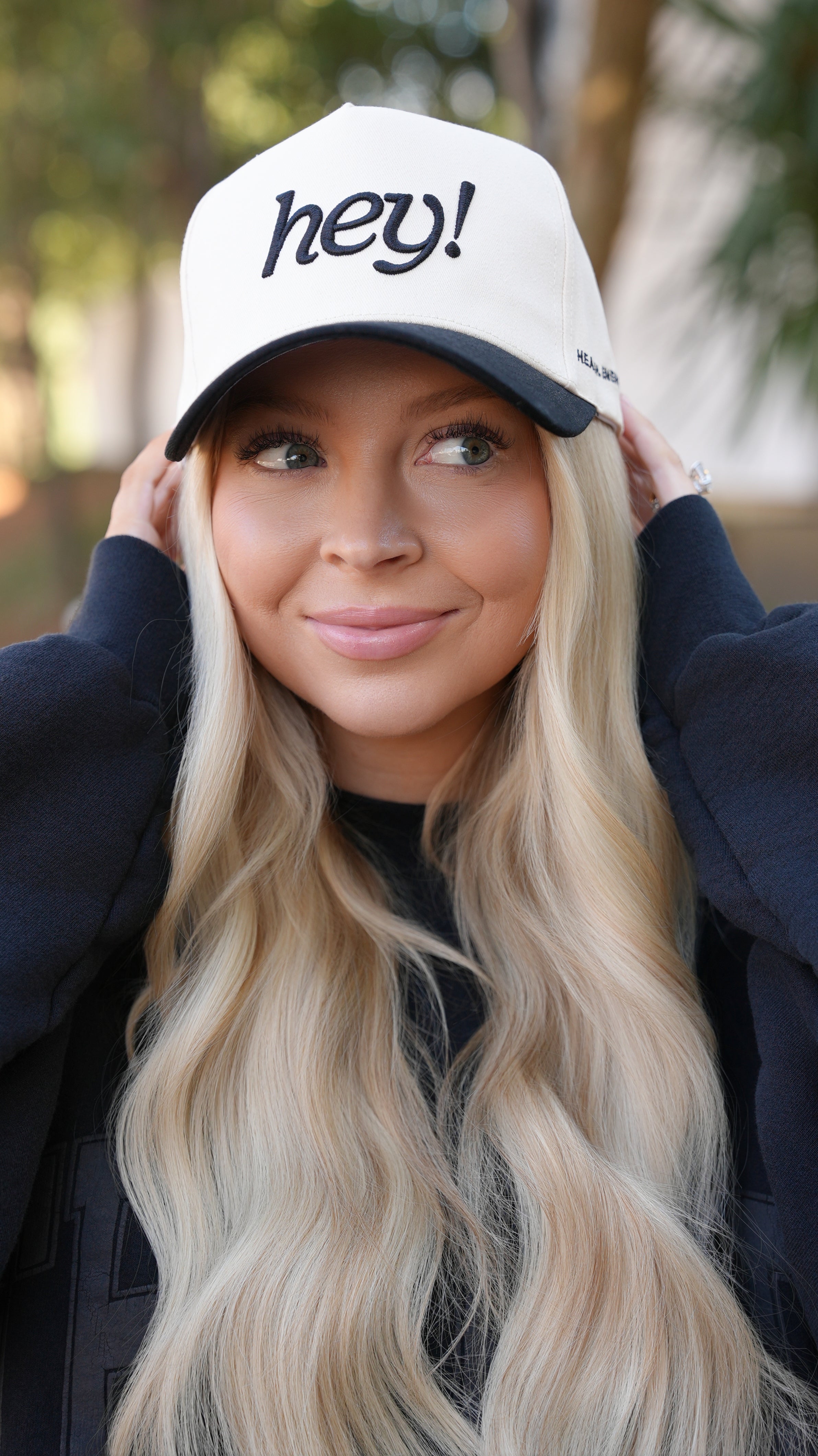 Kay Dudley wearing a cream “hey!” embroidered hat outdoors, smiling while adjusting the brim with both hands. 
