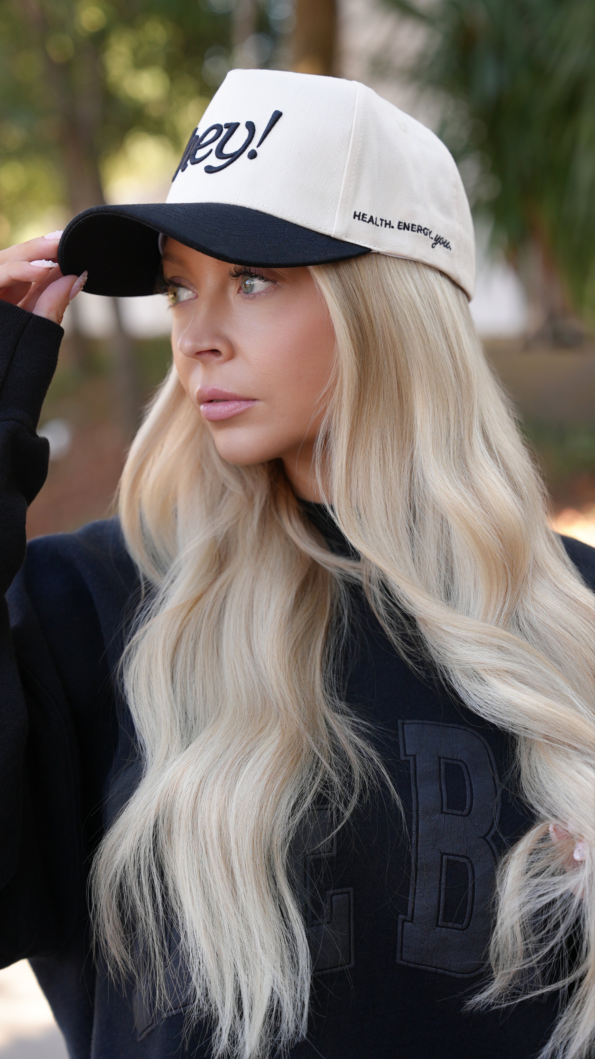 Kay Dudley, a blonde woman, wearing a black “hey!” embroidered hat outdoors, looking down while adjusting the brim.