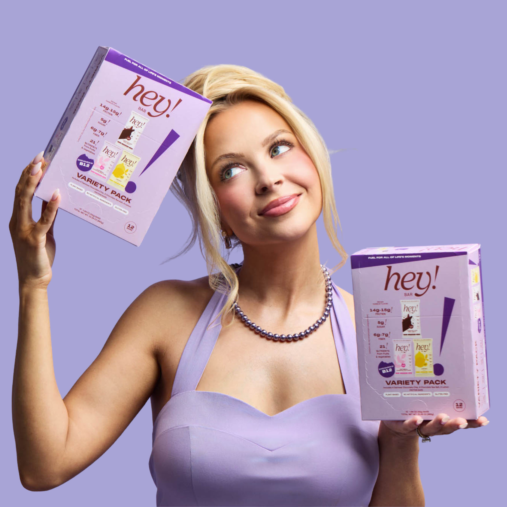Kay Dudley, a blonde woman, in a lavender dress holding two “hey!” variety pack protein bar boxes against a light purple background.