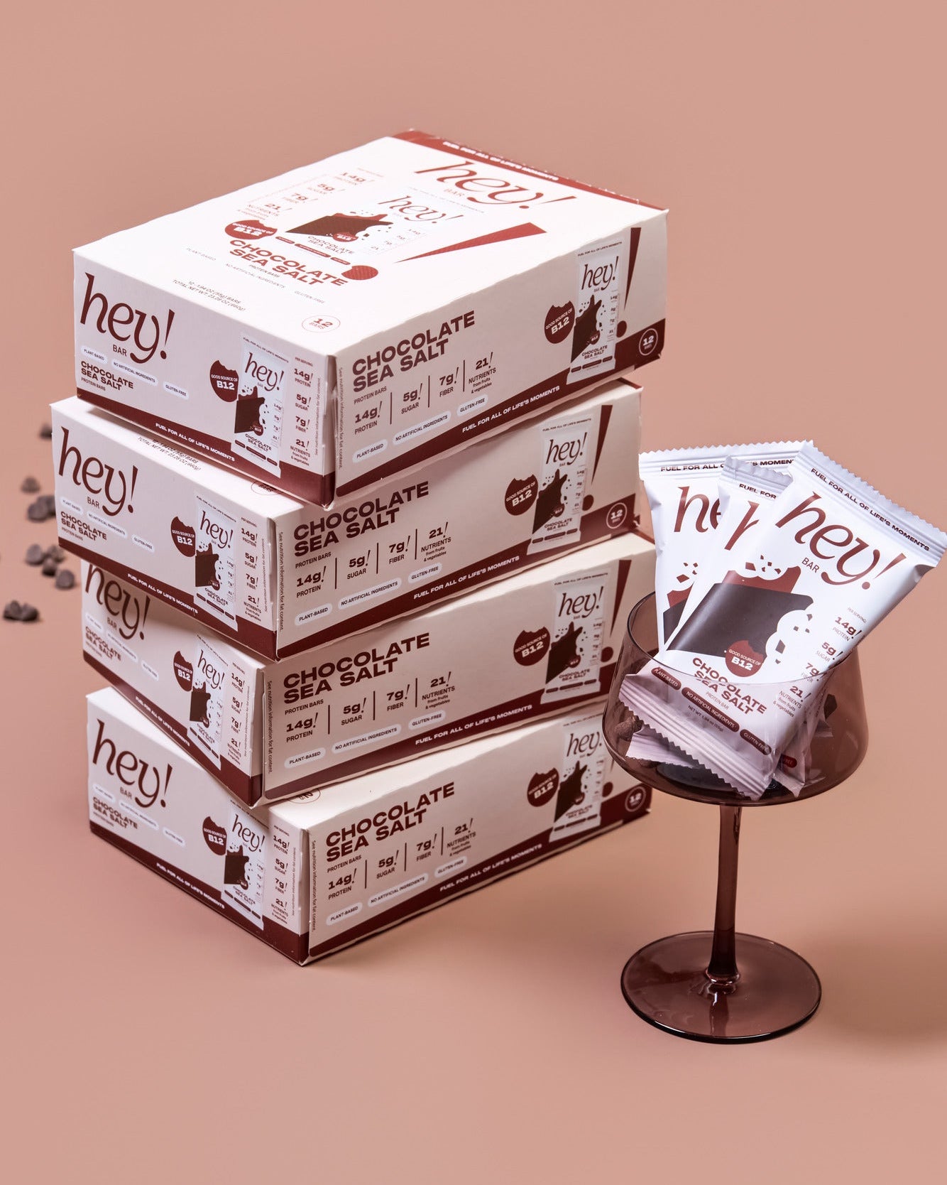 Four stacked HEY! BAR Chocolate Sea Salt cartons next to a glass dish holding three wrapped bars, on a warm brown background. 