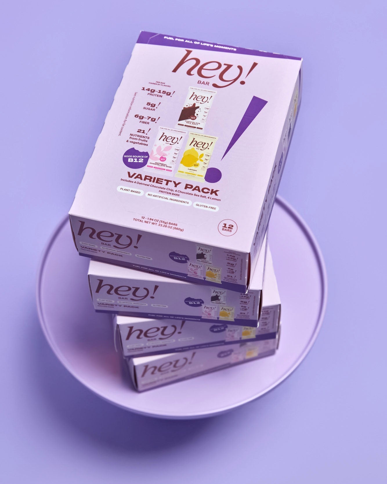 Four stacked HEY! BAR Variety Pack boxes on a clear round stand against a light purple background. 
