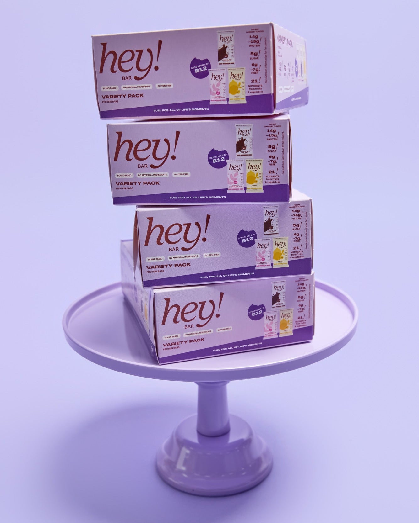 Four stacked HEY! BAR Variety Pack boxes on a clear round stand against a light purple background. 
