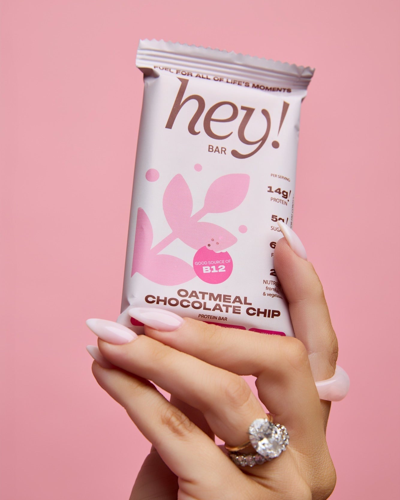 Hand with pale pink manicured nails and a large diamond ring holding a HEY! BAR Oatmeal Chocolate Chip wrapper (light lavender with pink leaf graphic and “hey!” logo) against a soft pink background.