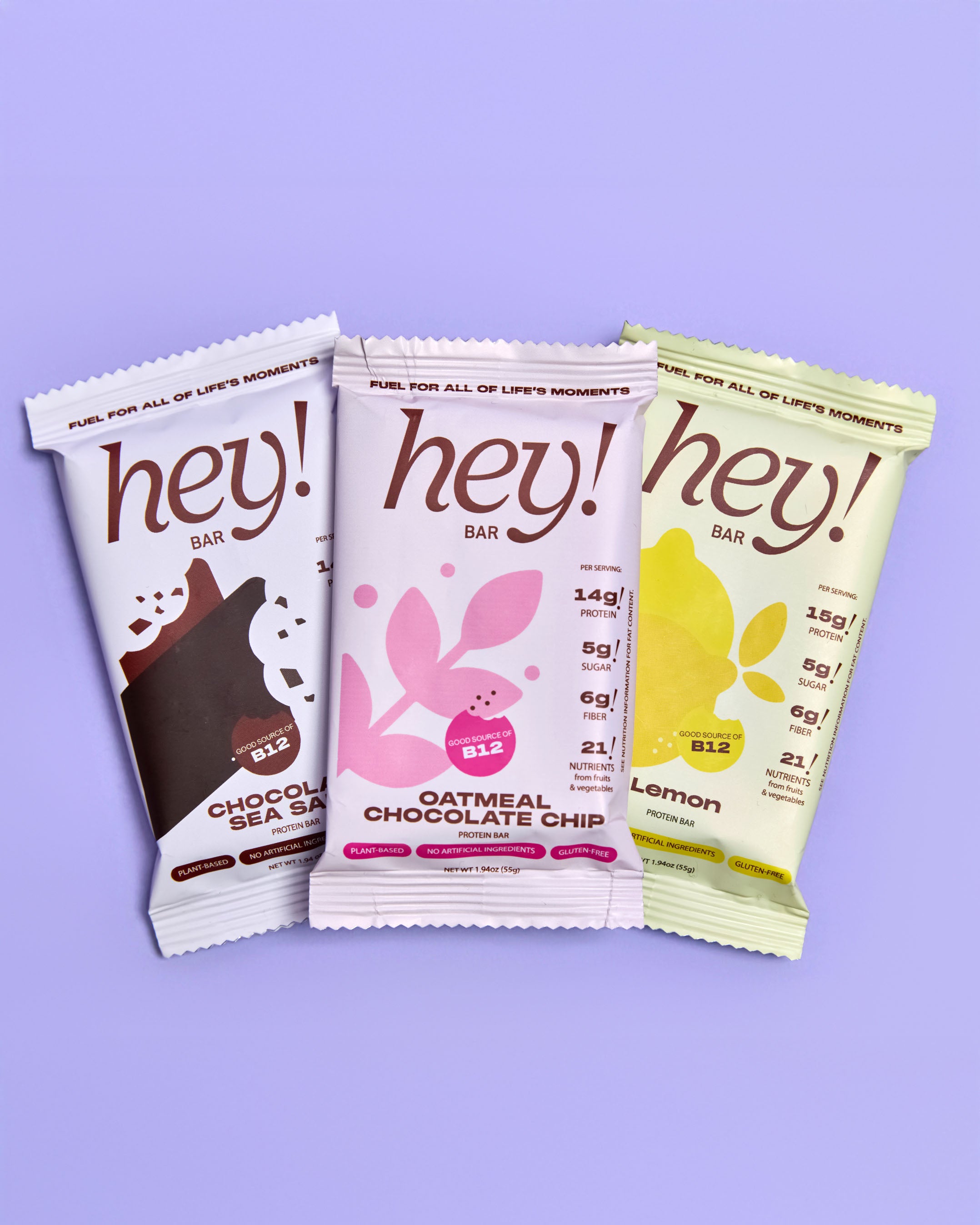 Three ‘hey!’ protein bar wrappers—Chocolate Sea Salt, Oatmeal Chocolate Chip, and Lemon—overlap on a light purple background.