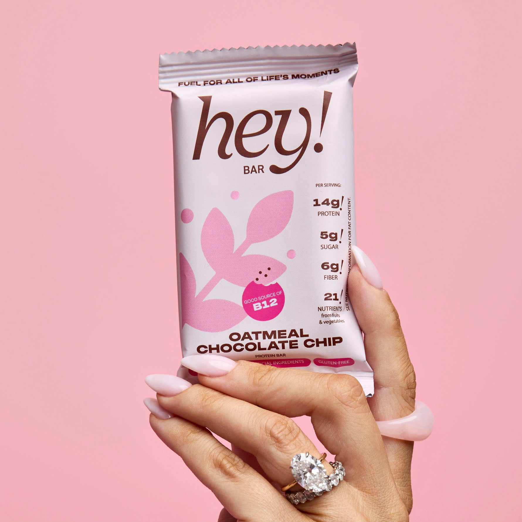 Hand with pink manicure and rings holds a ‘hey!’ Oatmeal Chocolate Chip protein bar wrapper against a pink background.