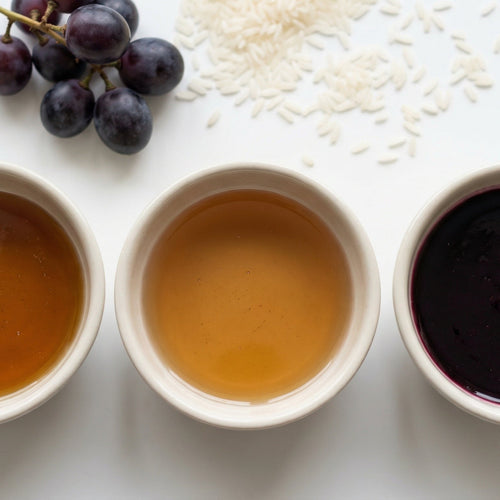 Three small bowls with different colored sugar syrups on a white surface with grapes and rice.
